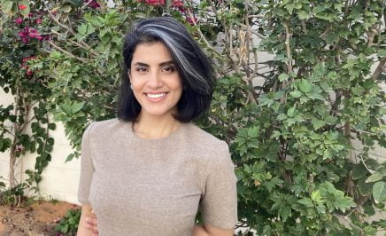 Loujain with black hair and a gray dress stands in front of a vine.