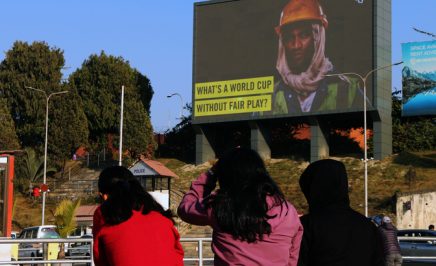 A group of young women in Qatar look at a billboard of a migrant worker with text 'What's a world cup without fair play?'