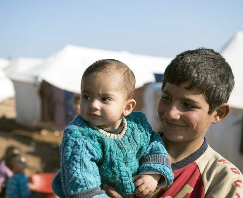 A smiling boy holds a baby, with white tents behind him