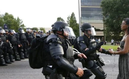 A black female demonstrator stands still in the street as US police officers in riot gear rush toward her. Behind them is a line of police officers also in riot gear.